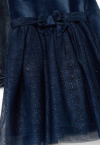 Abito navy con corpino di velluto, gonna in tulle con dettagli scintillanti e un fiocco abbinato in vita. Tessuto liscio con design stratificato.