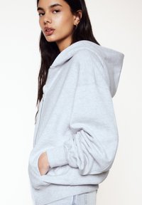 Grauer Zip-Hoodie mit weichem Material und lockerer Passform. Verfügt über eine Kapuze, große Vordertaschen und gerippte Bündchen. Kombiniert mit hellen Jeans.