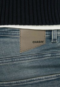 Maglione a coste scuro con orlo sopra un tessuto chiaro e jeans di denim blu con un'etichetta in pelle beige con la scritta "CHASIN'" cucita.