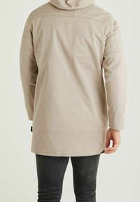 Beige långärmad jacka med huva som bärs av en person som står med ryggen mot kameran, i kombination med mörka slim-fit jeans.