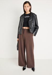 Μαύρο δερμάτινο cropped μπουφάν, εφαρμοστό μαύρο τοπ και καφέ παντελόνι με φαρδιά πόδια. Το μοντέλο κρατάει μια μαύρη τσάντα με ασημένιους τρουκς. Κοφτά μαύρα παπούτσια.