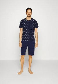 Tengerészkék rövid ujjú póló kis fehér polo logókkal, tengerészkék rövidnadrággal párosítva, puha anyagból készült. A modell mezítláb áll, semleges háttér előtt.