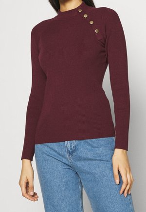 Femme portant un pull côtelé moulant bordeaux avec quatre boutons dorés sur une épaule et un jean bleu clair.