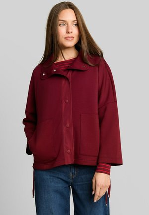 Vrouw met lang steil haar die een donkerrood jasje met knopen draagt over een gestreept shirt en een blauwe spijkerbroek, staand tegen een lichtgrijze achtergrond.