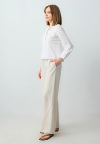 Witte blouse van oogjes met een kraag, lange mouwen en diamantpatronen. Gecombineerd met beige, wijde broeken en bruine slippers.
