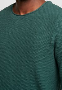 Zielony sweter o fakturze, z okrągłym dekoltem, posiadający wzór na tkaninie i ściągacz wokół kołnierza oraz szwów na ramionach.