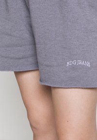 Šedé bavlněné šortky s volným střihem a nenastříhaným lemem, s bílým vyšitým nápisem "BDG JEANS" na levé noze.