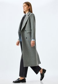Lang grå uldfrakke med klappet krave, dobbeltradede knapper, strukturerede skuldre og sidelommer, parret med mørkeblå jeans og sorte loafers.