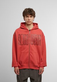 Coral zip-up hoodie with large "ROCA WEAR" logo. Features a hood, front pockets, and loose fit. Fabric appears soft and textured.