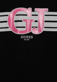T-shirt noir avec un logo "GJ" rose brillant, des rayures horizontales blanches et le texte "GUESS EST.1981" en blanc sous le logo. Texture de tissu douce.