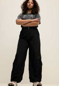 Zwarte cargobroek met een elastische tailleband, twee grote zijzakken en een gladde, lichte stof. Gecombineerd met een grijze T-shirt met een opdruk.