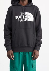 Persona che indossa un hoodie nero con il logo bianco "The North Face" e pantaloni cargo verdi su uno sfondo bianco uniforme.