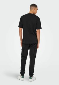 Camiseta de algodón negra y pantalones de carga negros, con bolsillos laterales y puños elásticos. El modelo está de espaldas, llevando zapatillas blancas.