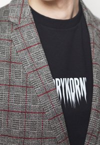 Grijze geruite blazer met rode accenten, gemaakt van een textuurstof. Daaronder een zwart shirt met vetgedrukt witte tekst.