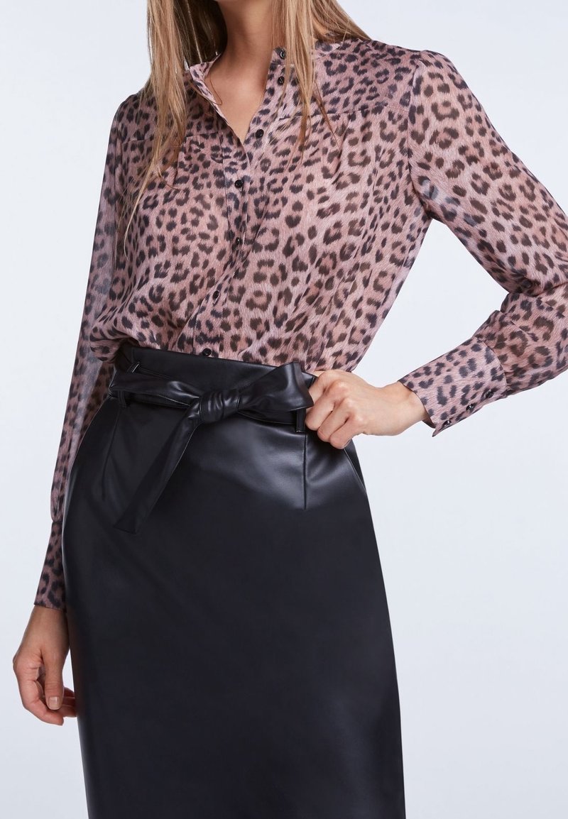 Une femme porte une blouse transparente à imprimé léopard rentrée dans une jupe en cuir noire taille haute, avec une ceinture nouée à la taille.