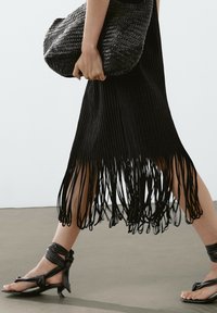 Svart plisserad klänning med fransar i nederkant, ihopparad med svarta sandaletter med remmar. Väskan är vävd och rundad, vilket tillför textur till outfiten.