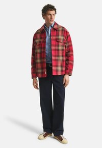 Rode geruite overhemdjas met twee borstzakken, gelaagd over een blauwe button-up shirt, gecombineerd met marineblauwe broek en bruine sneakers met witte zolen.