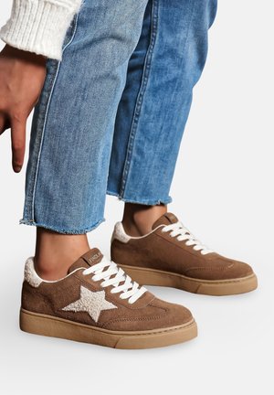 Baskets en suede marron avec un accent étoile blanc, un col texturé crème, des lacets blancs et une semelle en caoutchouc beige, portées avec un jean bleu délavé.