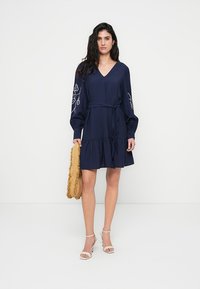 Μακρυμάνικο φόρεμα σε ναυτικό μπλε με V-λαιμόκοψη και φουσκωτά μανίκια, διακοσμημένο με κεντήματα με λουλούδια στους μανσέτες. Διαθέτει ζώνη στη μέση και βολάν με σκάλες στο τελείωμα. Φοράται με μπεζ παπούτσια.