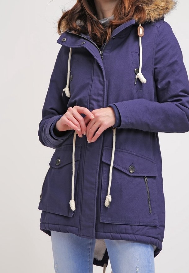 Personne portant une parka bleue avec des cordons blancs, une capuche doublée de fourrure, des poches à boutons et un jean bleu clair, debout les mains jointes.