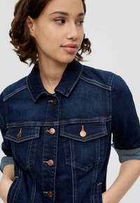 Donkerblauwe spijkerjas met metalen knopen, twee borstzakken, opgerolde mouwen en zichtbare stiksels.