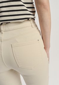 Beżowe dżinsy stretch z detalami na tylnych kieszeniach i kontrastowymi szwami, noszone z czarną i białą pasiastą bluzką.