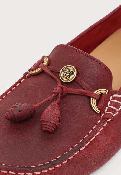 Mocassin en daim rouge avec couture blanche, pampilles en cuir décoratives et un emblème doré de Méduse à l'avant supérieur.