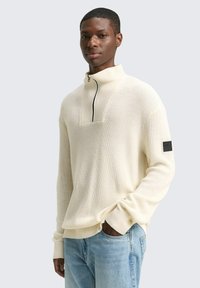 Cremfarbener Strickpullover mit hohem Kragen und Viertel-Reißverschluss, mit quadratischem Ausschnitt und einem schwarzen Stoffetikett am Ärmel.