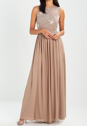 Ärmelloses beiges Abendkleid mit transparentem, verziertem Oberteil und fließendem, plissiertem Rock, kombiniert mit silbernen High-Heel-Sandalen.