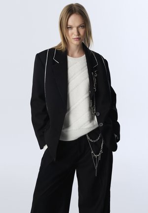 Zwarte op maat gemaakte blazer met witte piping, gelaagd over een witte gestructureerde sweater, gecombineerd met zwarte wijde broek met kettingaccenten.