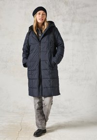 Abrigo largo azul marino acolchado con capucha, cierre de cremallera, bolsillos frontales y tejido texturizado; llevado sobre una camisa a cuadros y pantalones grises, combinados con zapatos negros.