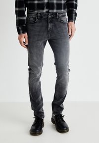 Chemise à carreaux noir et gris à manches longues, assortie à un jean noir délavé et des chaussures noires avec des coutures jaunes.