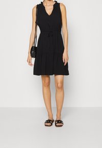 Svart ärmlös klänning med V-ringning och volangdetaljer, åtstramad midja, flerlagad kjol och tillsammans med svarta halkskor.