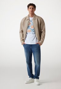 Beige bomberjack, witte t-shirt met grafische bloemenontwerp, blauwe spijkerbroek, witte sneakers, staand tegen een lichte achtergrond.
