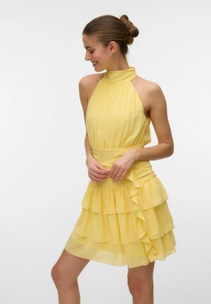 Femme en robe jaune sans manches à col halter avec jupe à volants superposés, souriante et regardant vers sa gauche sur un fond blanc uni.