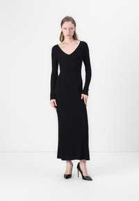 Black ribbed knit maxi dress with long sleeves and a V-neck. Smooth texture, fitted silhouette, and flared hem. Worn with black high heels.