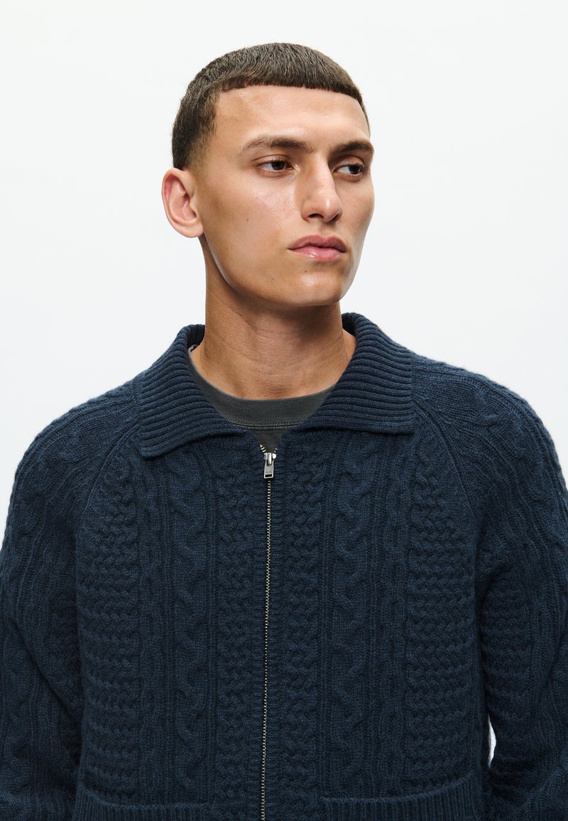 Navy-blauer Sweater aus Kabelstrick mit hohem Kragen und Reißverschluss. Verfügt über strukturierte Muster und einen mittellangen Schnitt. Weiches, warmes Material.