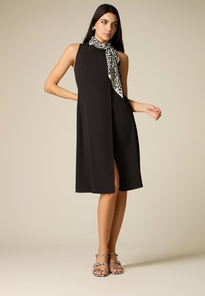 Donna con lunghi capelli scuri che indossa un vestito nero senza maniche con spacco frontale, sciarpa leopardata annodata al collo e sandali coordinati con stampa leopardata.