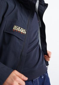 Napa herrejakke i marineblå, med lynlåsfortøjning og logo patch. Har et struktureret stof, stående krave og elastiske manchetter. Lagd ovenpå en mørk skjorte.