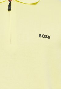 Rumena polo majica na zadrgo z rebrasto teksturo, z značko "BOSS" v črni barvi na levi strani. Zadrga na ovratniku.