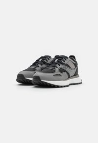 Chaussures de sport grises et noires présentant un mélange de matériaux lisses et texturés, avec une semelle blanche épaisse et des accents de logo bien visibles.