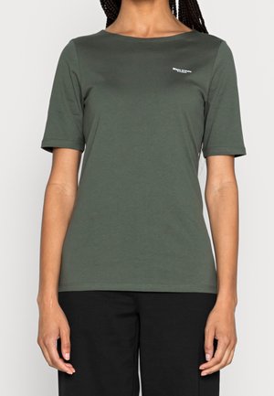 Mujer con camiseta de manga corta de color verde oliva con pequeño texto blanco en la parte superior izquierda y pantalones negros, de pie frente a un fondo sencillo.
