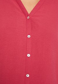 Camiseta vermelha de manga curta com decote em V e textura suave. Apresenta botões brancos detalhando a pala frontal, com um pequeno recorte acima dos botões.