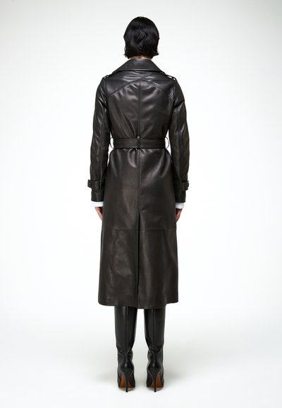 Personne vue de dos portant un long trench-coat en cuir noir avec ceinture et des bottes noires à talons hauts sur un fond blanc.