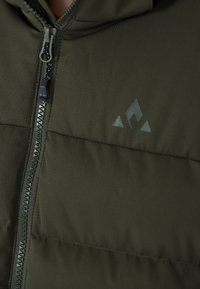 Grön vadderad jacka med dragkedja, med en textur med svaga linjer och en mörk logotyp med triangulära detaljer på bröstet.