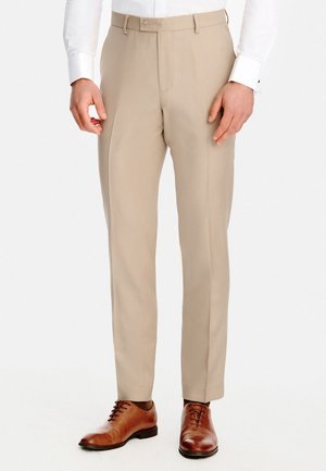 Mężczyzna w beżowych obcisłych spodniach, białej koszuli wizytowej i brązowych skórzanych eleganckich butach, stojący na jednolitym tle.