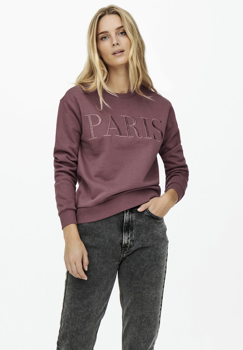 Κά sweatshirt σε καφέ χρώμα με την ένδειξη "ΠΑΡΙΣ" σε ανάγλυφα γράμματα. Μακριά μανίκια, στρογγυλή λαιμόκοψη και ριμπ μανσέτες. Φοριέται με ξεθωριασμένο μαύρο τζιν.