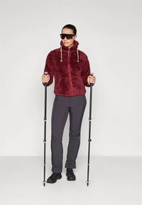 Bordo spalvos fliso striukė su užtrauktuku ir kapišonu, derinama su pilkomis kelnėmis ir juodais žygių batais. Laiko žygio lazdas, dėvi akinius nuo saulės.