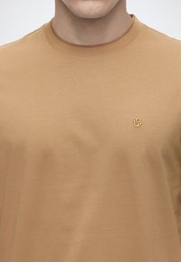 Άνδρας που φοράει ένα απλό ανοιχτό καφέ T-shirt με στρογγυλή λαιμόκοψη και μικρό κεντημένο λογότυπο στο αριστερό μέρος του στήθους.