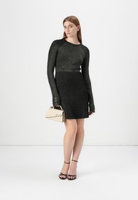 Modella in posa con un vestito nero brillantini a maniche lunghe, che tiene una borsa beige, indossando tacchi neri con cinturini, su uno sfondo bianco.